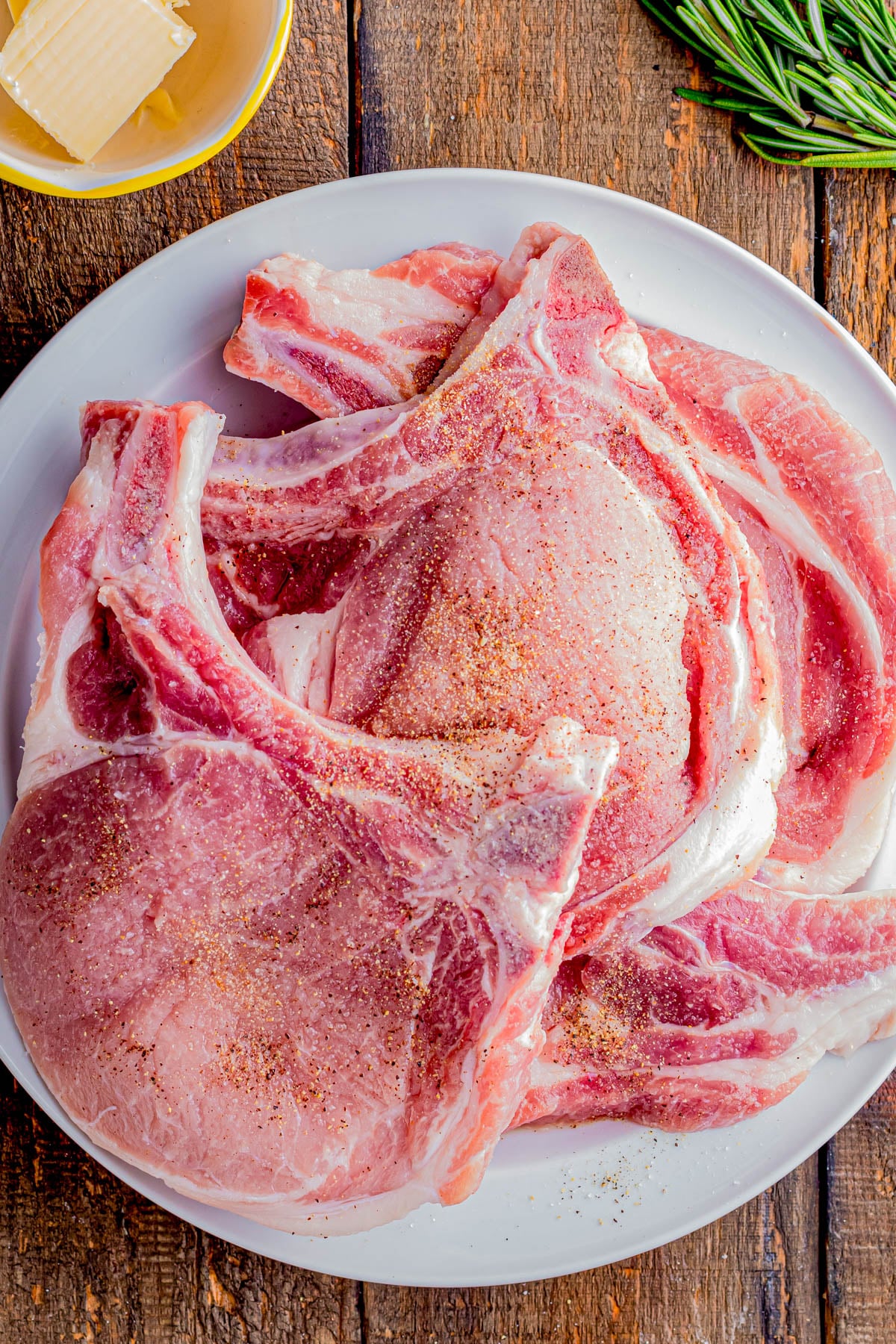 Raw bone-in pork chops seasoned with spices are arranged on a white plate, with a bowl of butter and a sprig of rosemary nearby on a wooden surface.