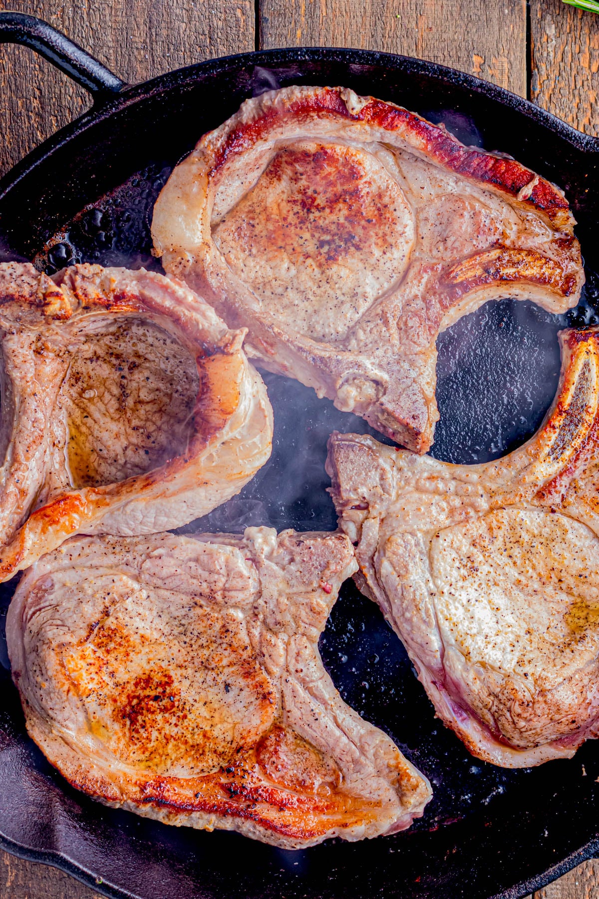 Four bone-in pork chops are being seared in a black cast iron skillet, with visible browning and steam rising from the hot pan.