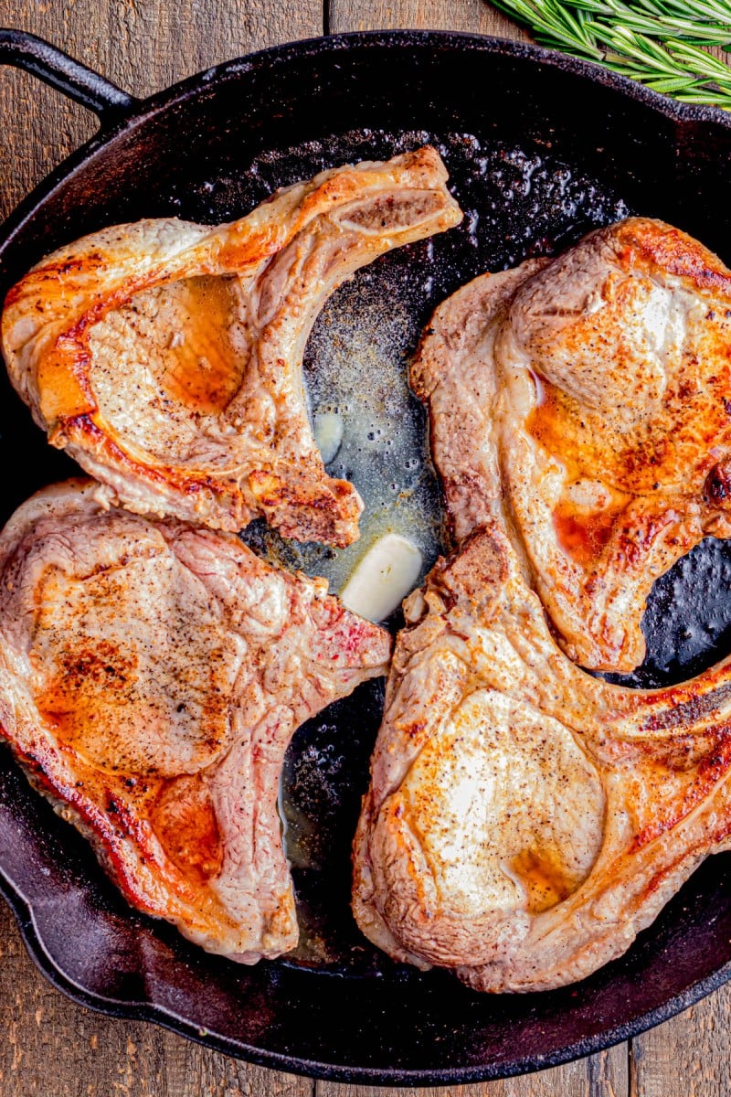 Four seasoned pork chops are being seared in a cast iron skillet with melted butter and garlic cloves.