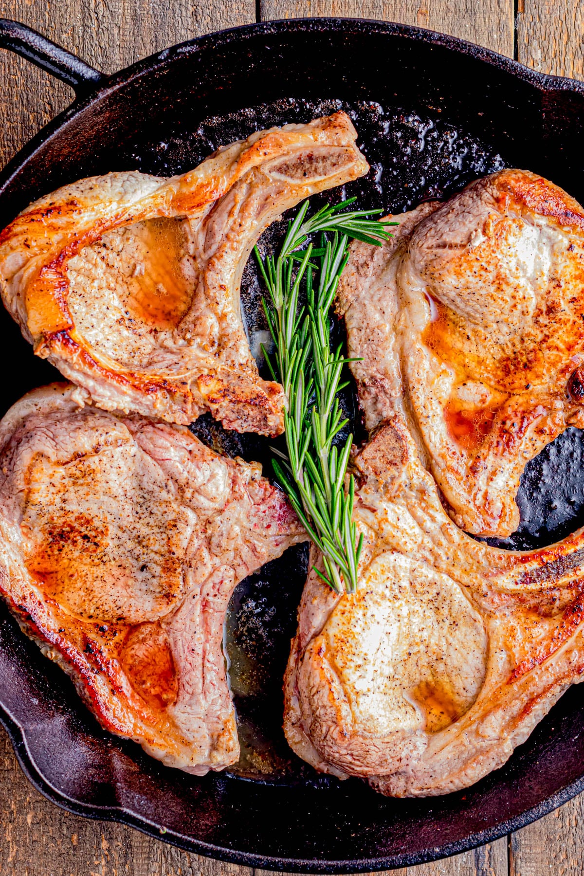 Four cooked pork chops with a sprig of rosemary in the center, arranged in a cast iron skillet on a wooden surface.