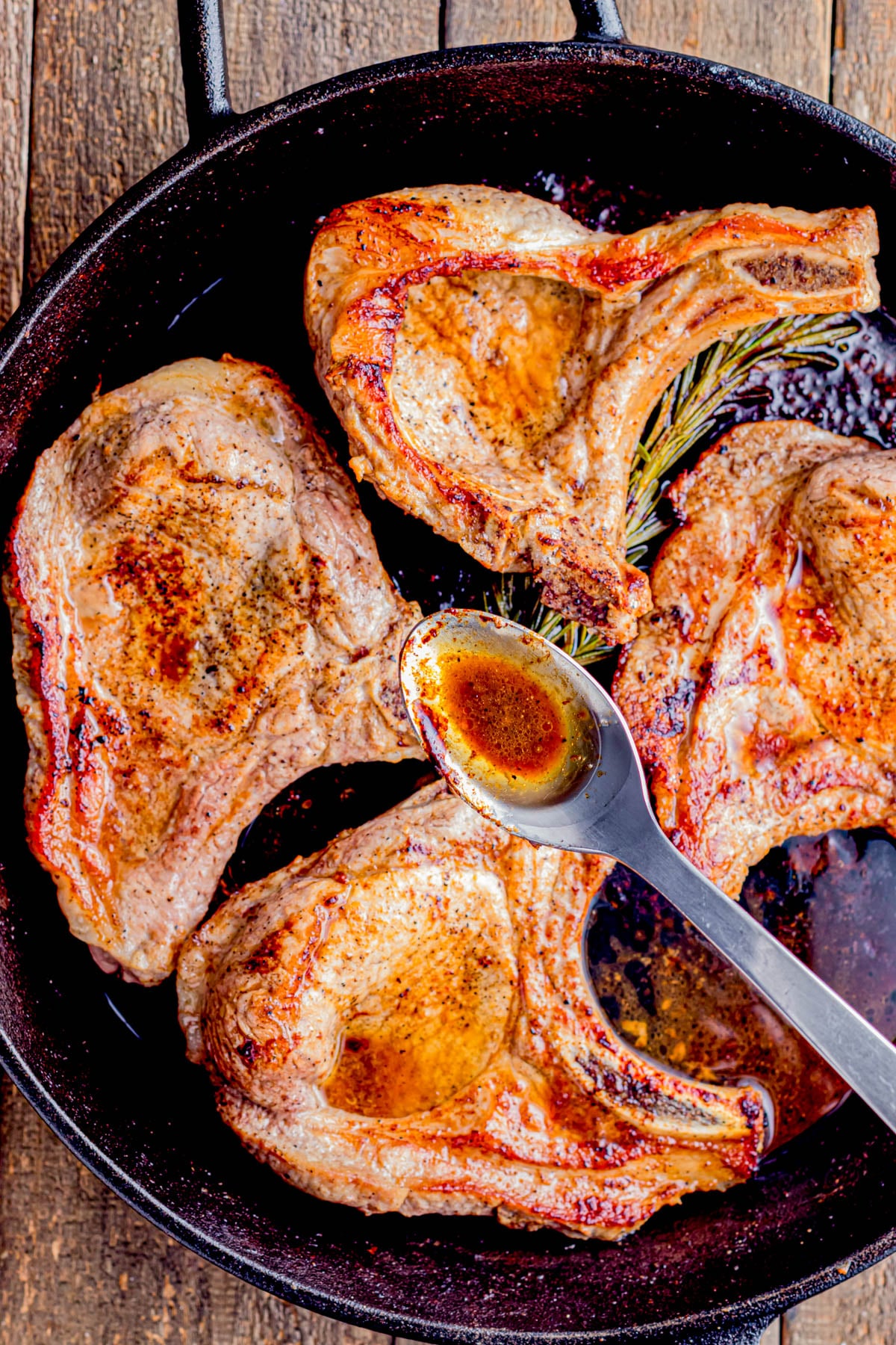 Three cooked pork chops in a cast iron skillet with a spoon drizzling sauce over one chop; a sprig of rosemary is also visible in the pan.