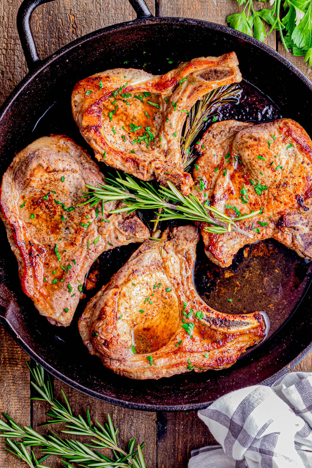 Four cooked pork chops garnished with chopped herbs and rosemary sprigs in a cast iron skillet, with parsley and a napkin nearby on a wooden surface.