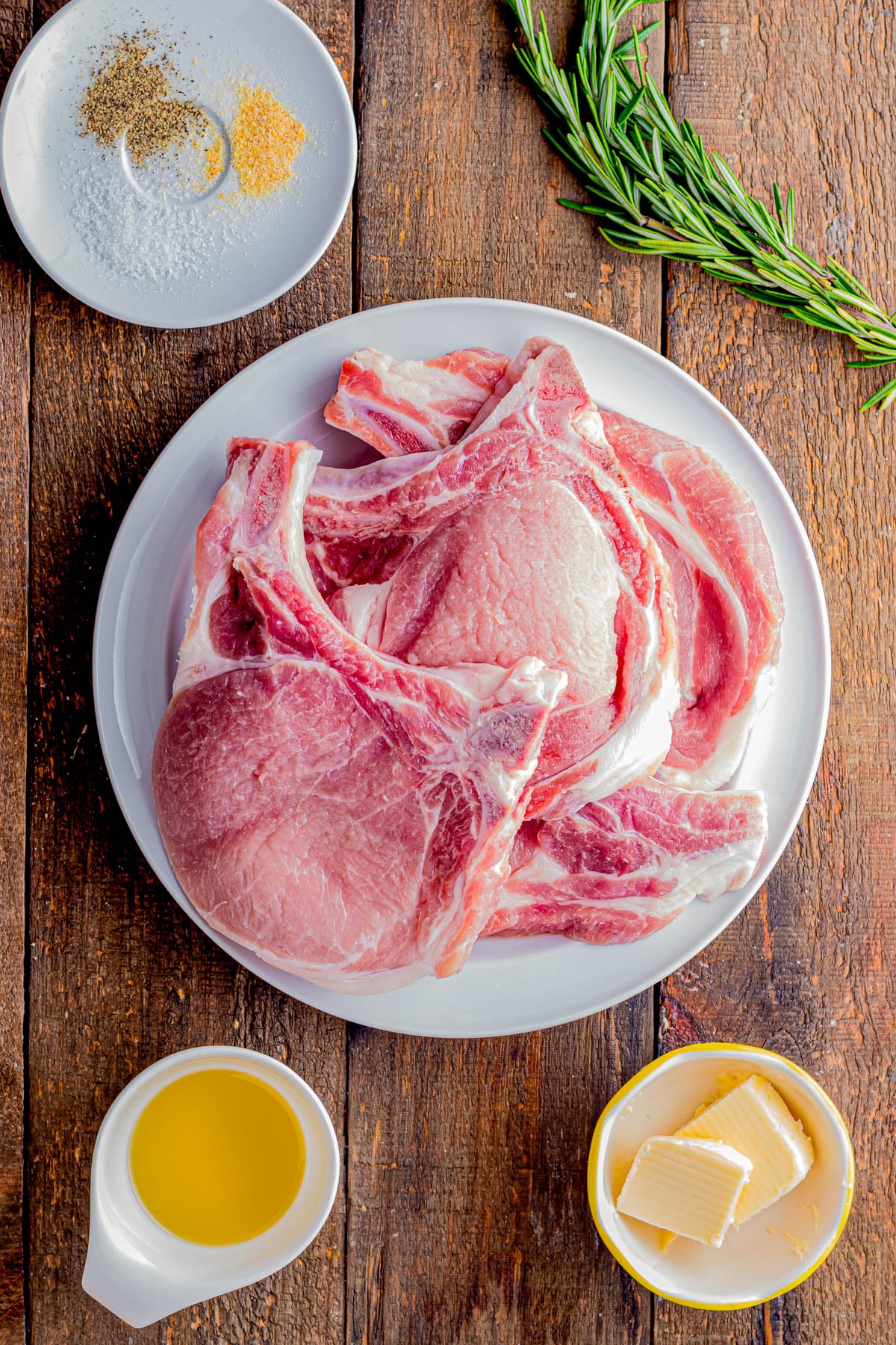 Raw pork chops on a white plate, with rosemary, a dish of salt and pepper, a bowl of olive oil, and a bowl of butter on a wooden surface.