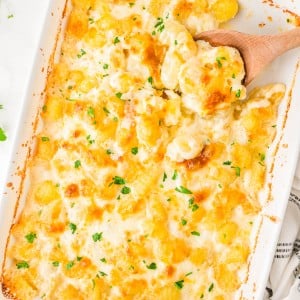 Baked cheesy potato casserole in a white dish, topped with melted cheese and chopped parsley, with a wooden spoon scooping a serving.