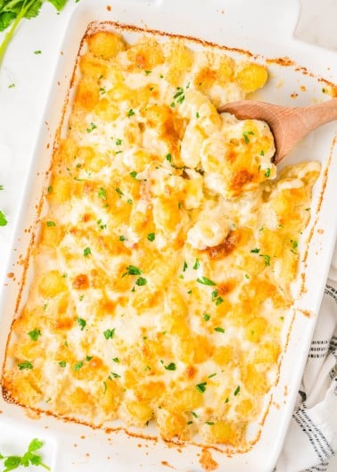 Baked cheesy potato casserole in a white dish, topped with melted cheese and chopped parsley, with a wooden spoon scooping a serving.