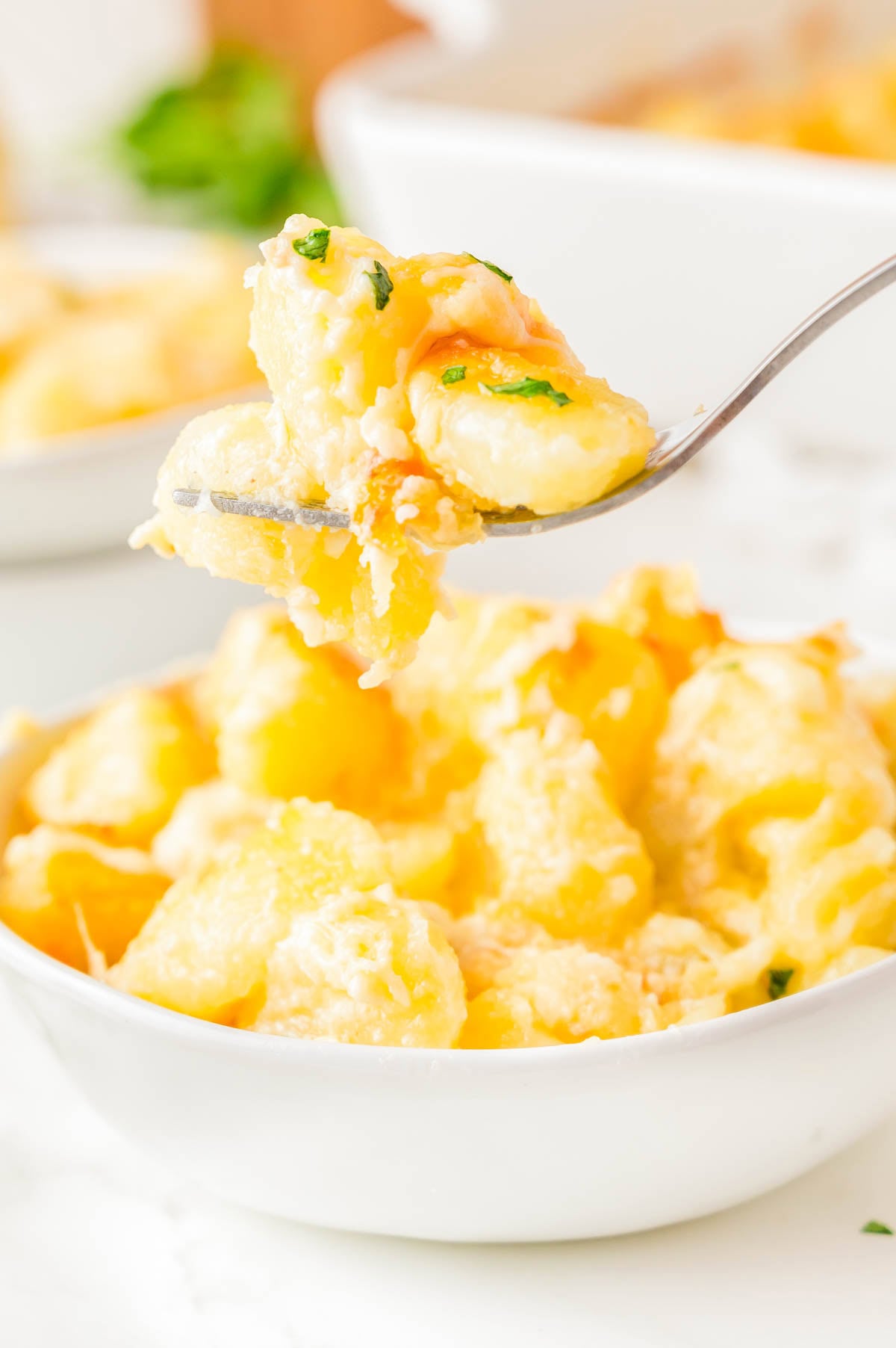 A close-up of a fork holding a bite of cheesy macaroni and cheese above a white bowl filled with more macaroni and cheese.