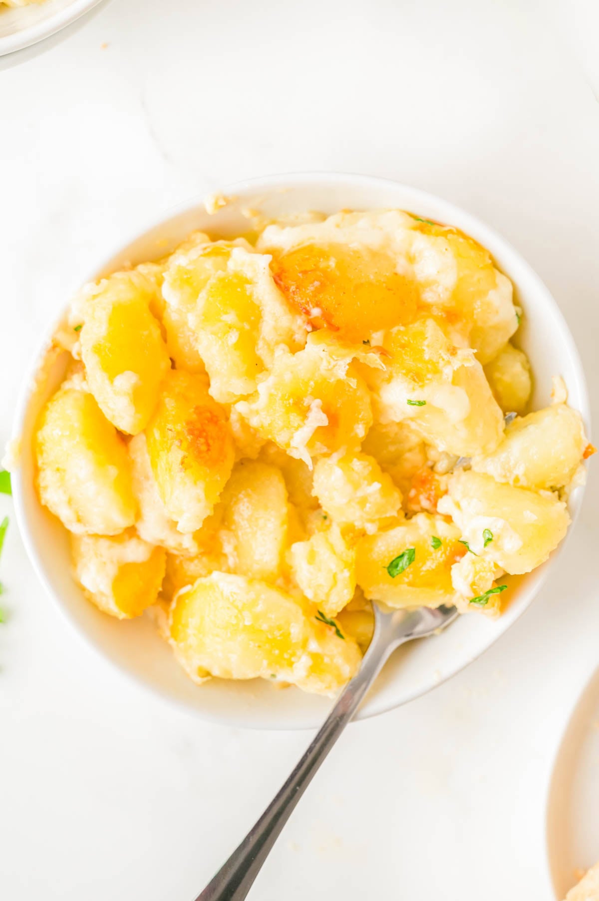 A bowl of cheesy baked gnocchi with a spoon, topped with melted cheese and garnished with herbs.