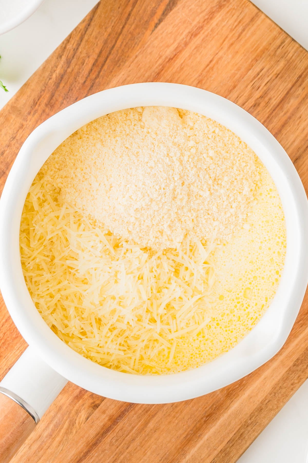 A white saucepan on a wooden board contains a yellow liquid topped with grated cheese and breadcrumbs.