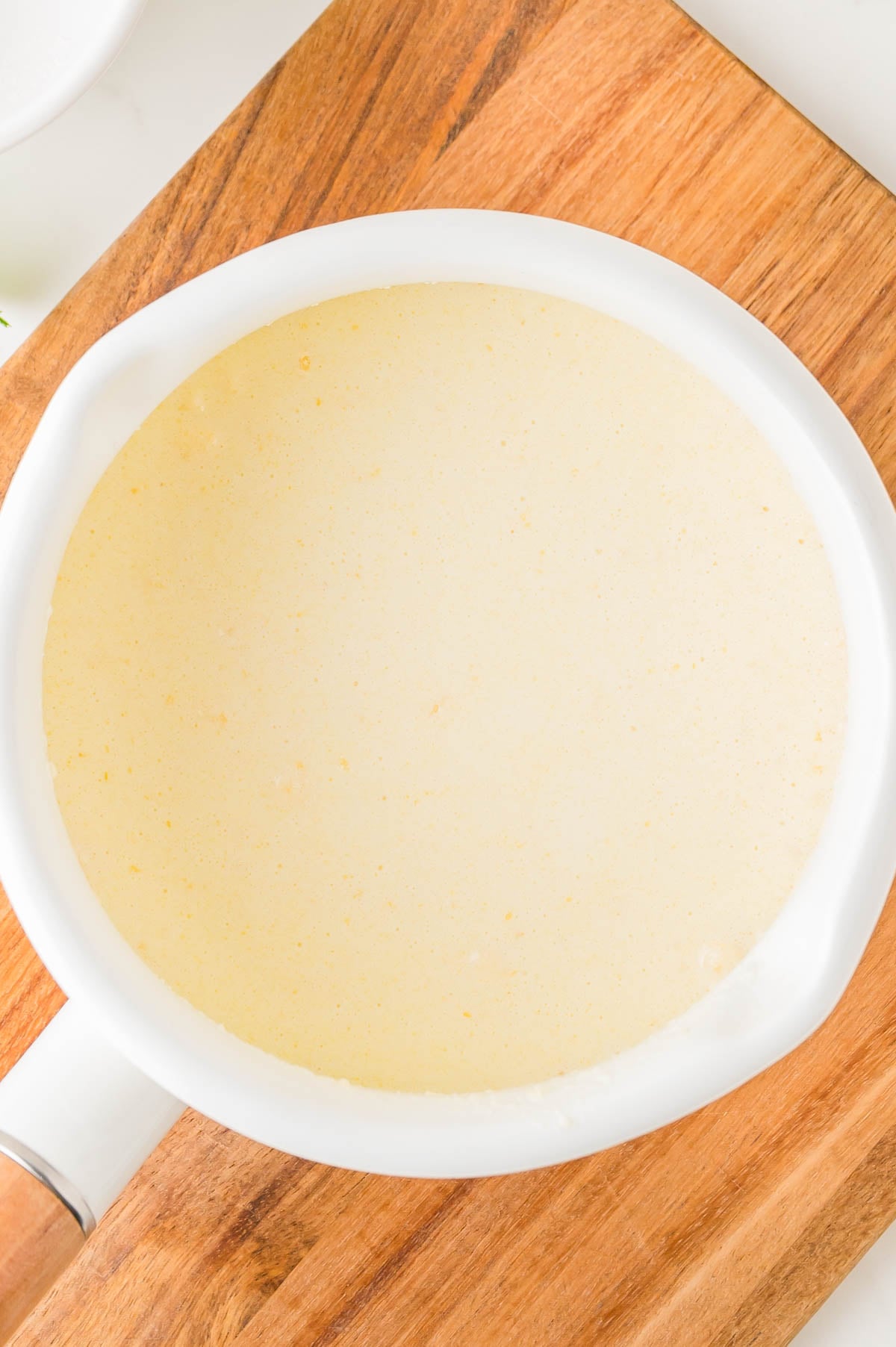 White saucepan filled with a light beige liquid, resting on a wooden cutting board viewed from above.