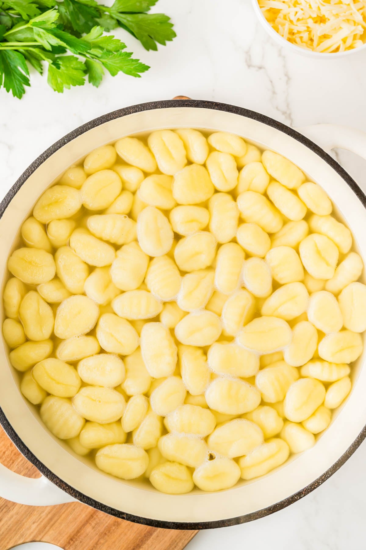 A pot filled with gnocchi boiling in water, surrounded by parsley and shredded cheese on a white countertop.