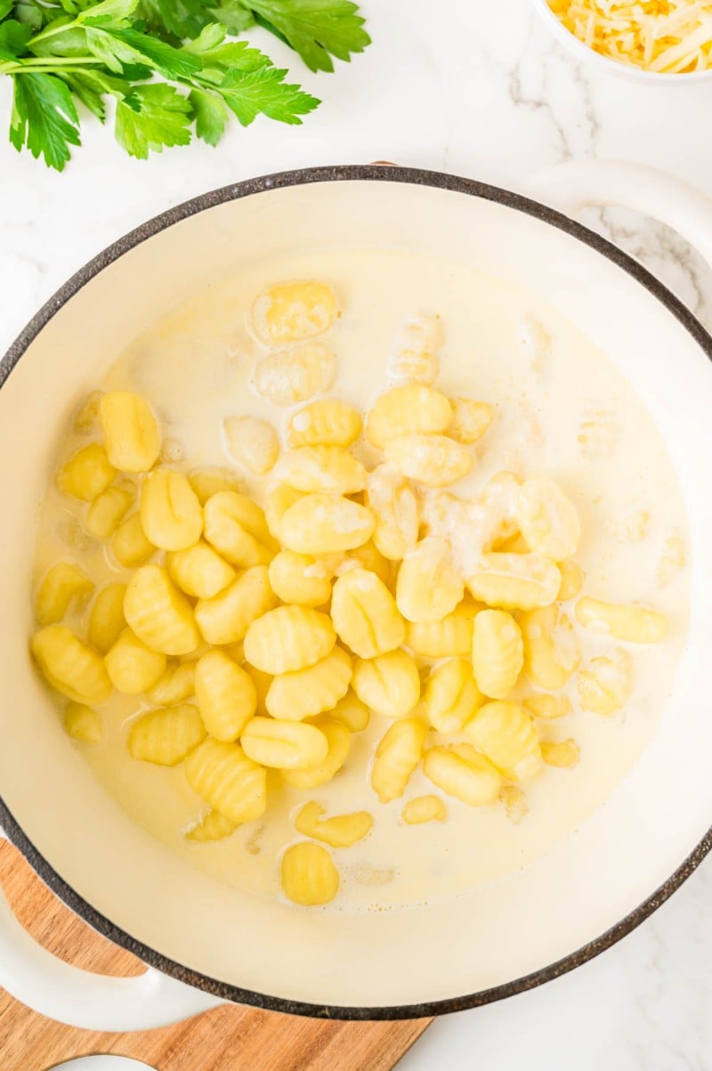 A white pot filled with gnocchi cooking in a creamy sauce, resting on a wooden board with parsley and grated cheese nearby.