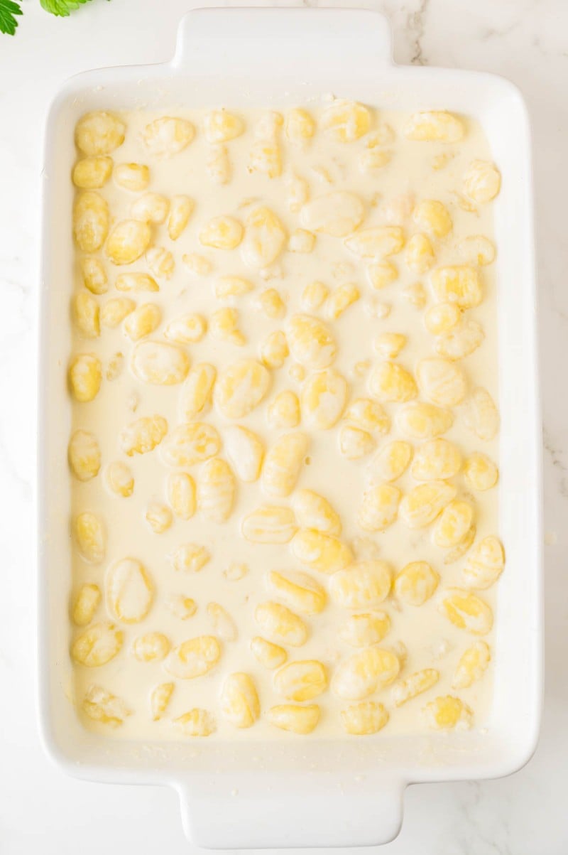 White baking dish filled with uncooked gnocchi covered in creamy white sauce, photographed from above on a light surface.