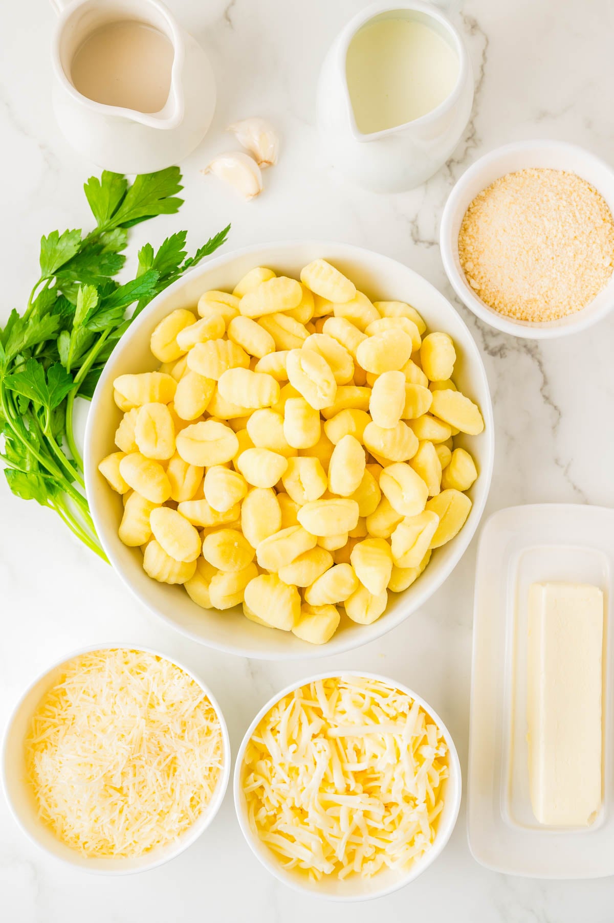 Bowl of uncooked gnocchi surrounded by fresh parsley, grated cheeses, breadcrumbs, butter, cream, and two garlic cloves on a white surface.
