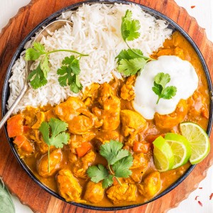 A plate of white rice and chicken curry garnished with cilantro, lime wedges, and a dollop of yogurt, served on a round wooden board with a fork.