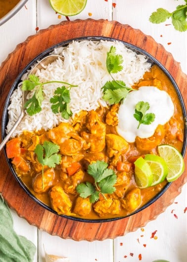 A plate of white rice and chicken curry garnished with cilantro, lime wedges, and a dollop of yogurt, served on a round wooden board with a fork.
