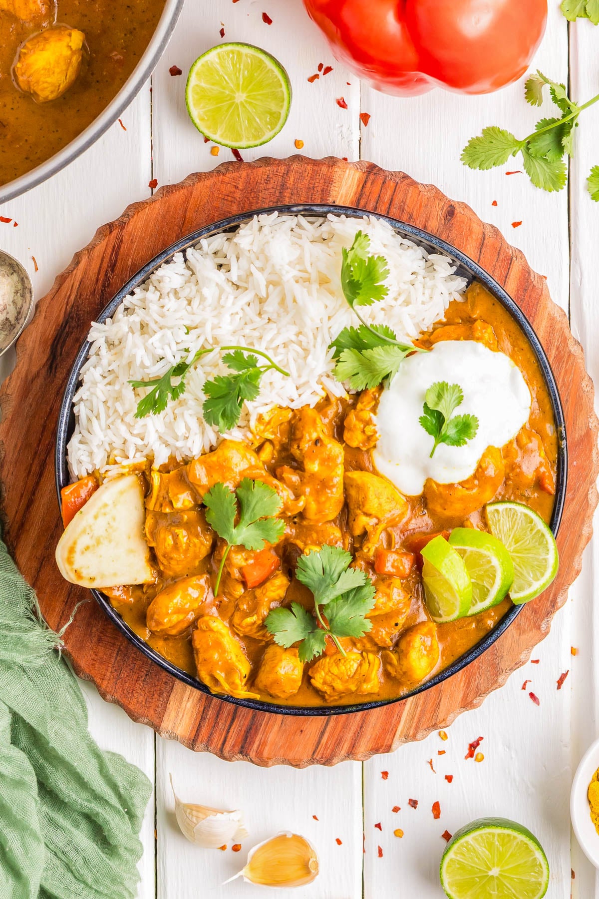 A bowl of white rice and chicken curry garnished with cilantro, lime wedges, and a dollop of yogurt, placed on a wooden board with ingredients around.