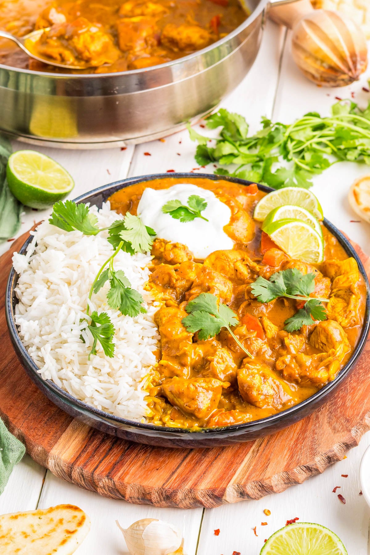 A plate with white rice, chicken curry topped with cilantro, lime slices, and a dollop of yogurt, served on a wooden board with naan and herbs nearby.