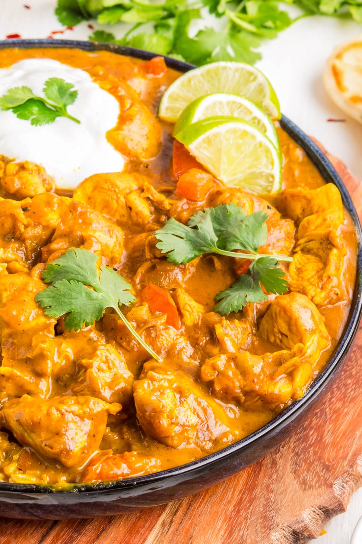 A bowl of chicken curry garnished with cilantro, served with lime slices and a side of yogurt, on a wooden surface.