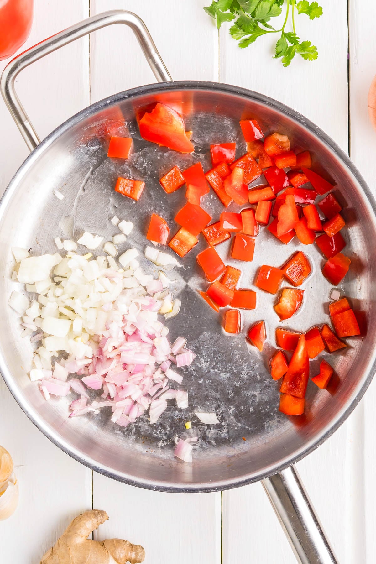 A stainless steel pan with chopped onions, shallots, and red bell peppers being sautéed, seen from above on a white wooden surface.