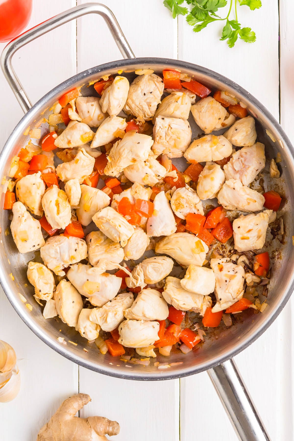 Chunks of cooked chicken breast, diced red bell pepper, and chopped onion in a stainless steel skillet on a white wooden surface.