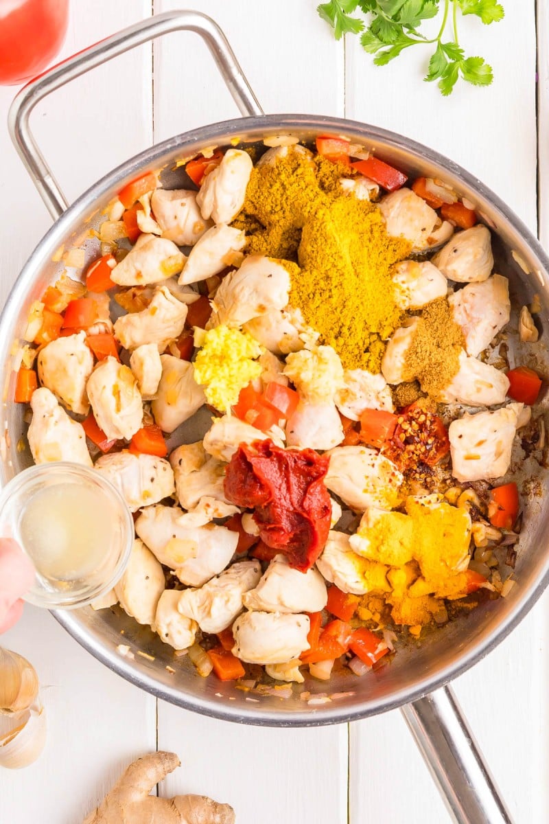 Chunks of chicken, diced red bell pepper, onions, and various spices cook in a stainless steel skillet; a hand pours liquid into the pan.