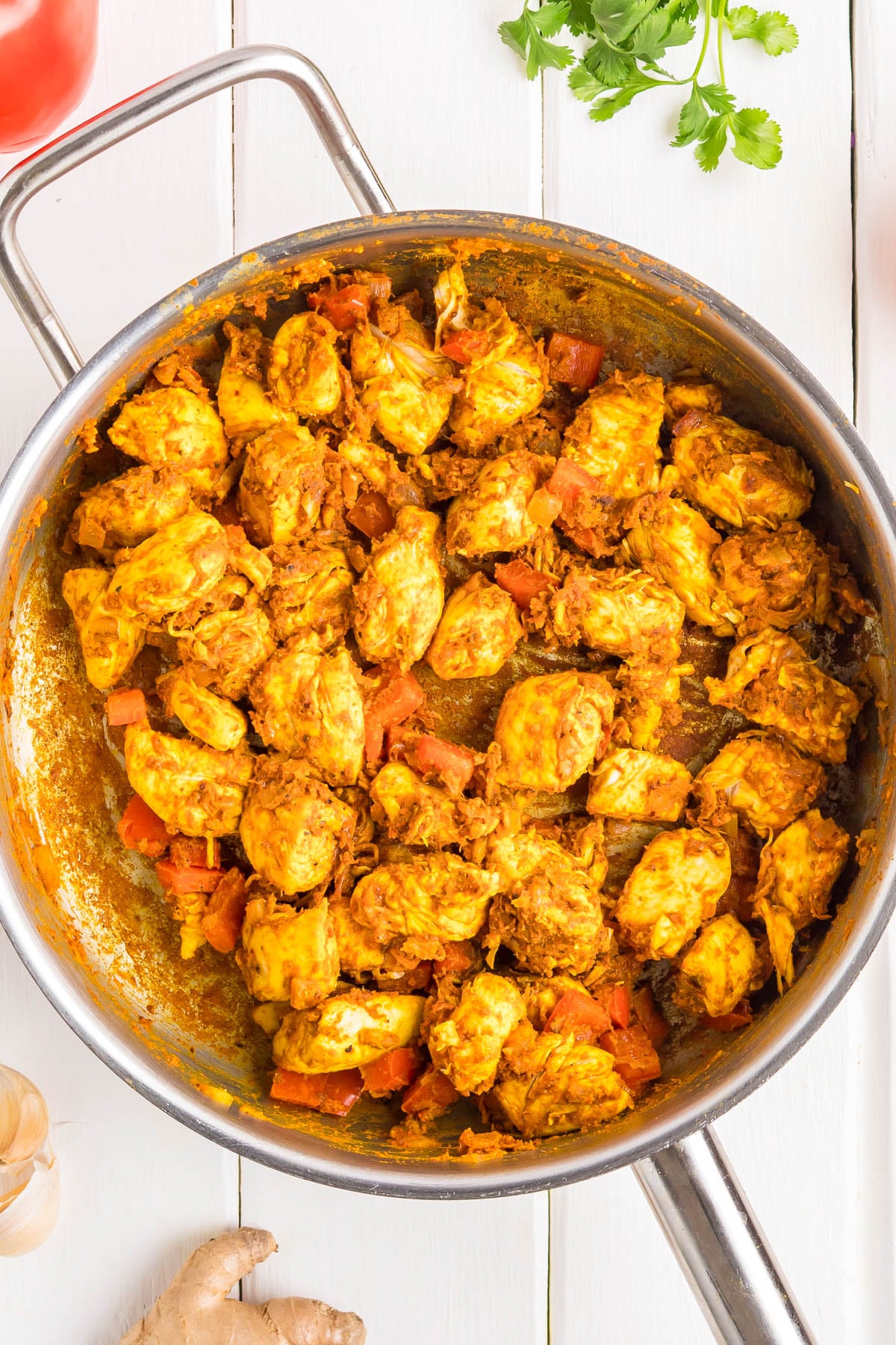 A stainless steel pan filled with cooked, cubed chicken pieces, sautéed with spices and small tomato chunks, sits on a white wooden surface.