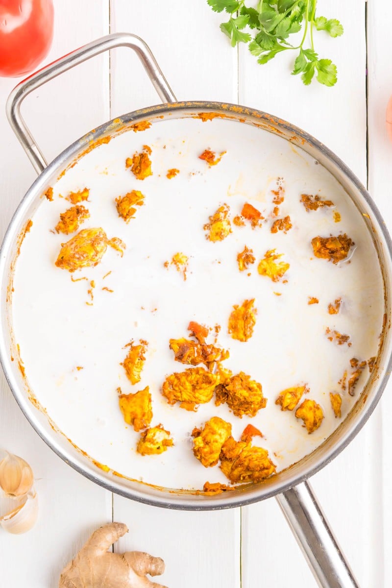 A stainless steel pan with pieces of cooked marinated chicken simmering in a creamy white sauce, set on a white wooden surface with ginger, garlic, tomato, and cilantro nearby.