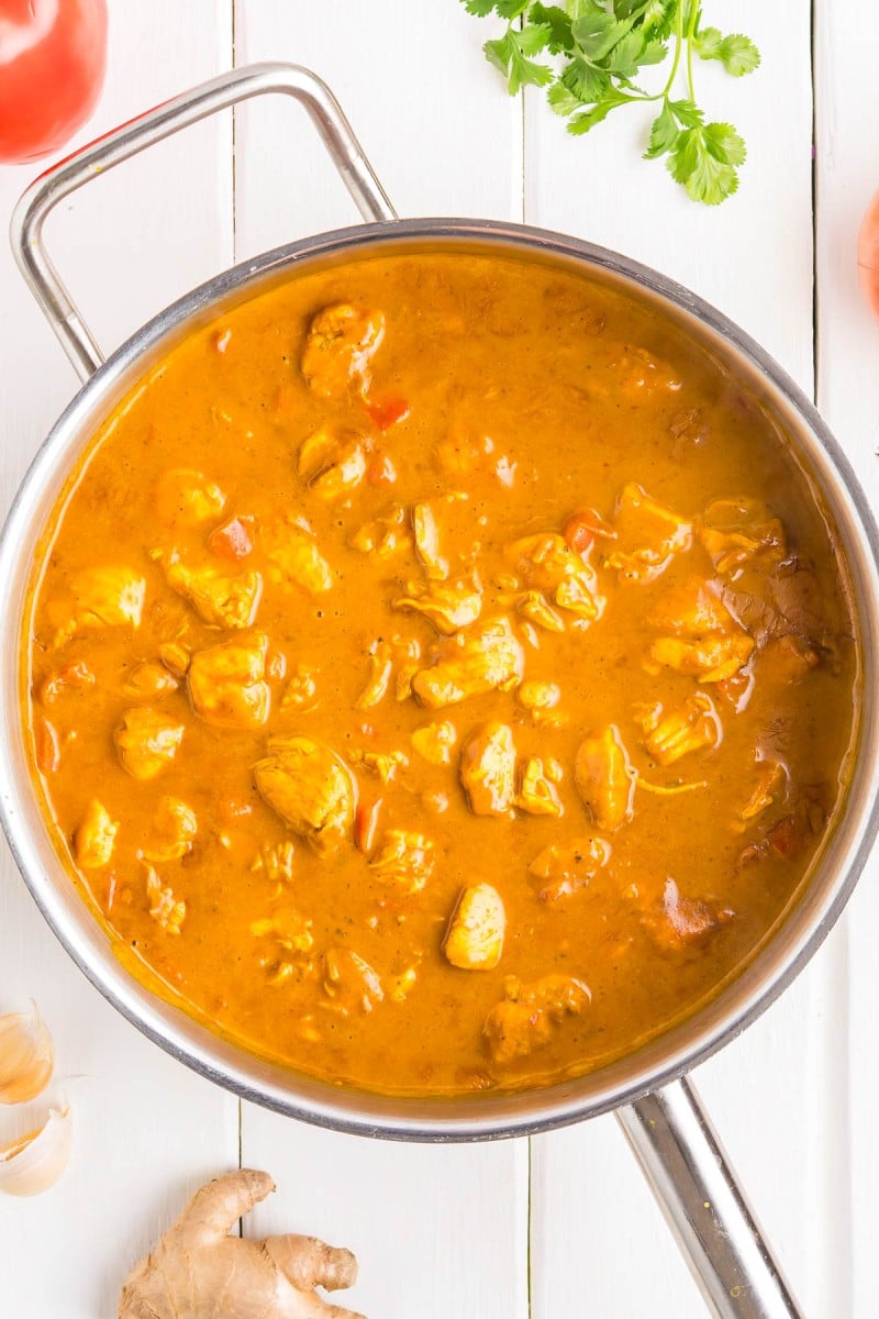 Stainless steel pan filled with orange curry containing chunks of meat, placed on a white surface with ginger, tomato, garlic, and cilantro nearby.