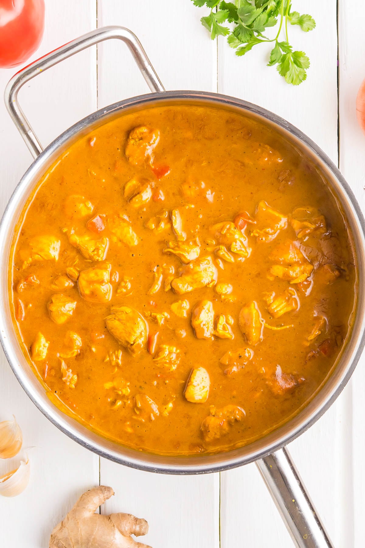 Stainless steel pan filled with orange curry containing chunks of meat, placed on a white surface with ginger, tomato, garlic, and cilantro nearby.