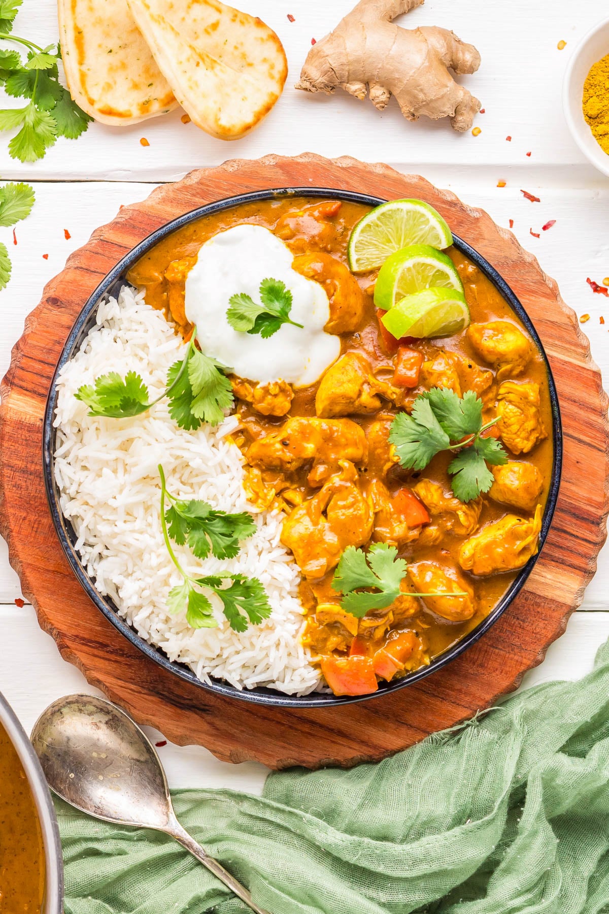 A bowl with white rice, chicken curry, cilantro, lime slices, and a dollop of yogurt, served on a wooden platter with naan bread, ginger, and spices nearby.