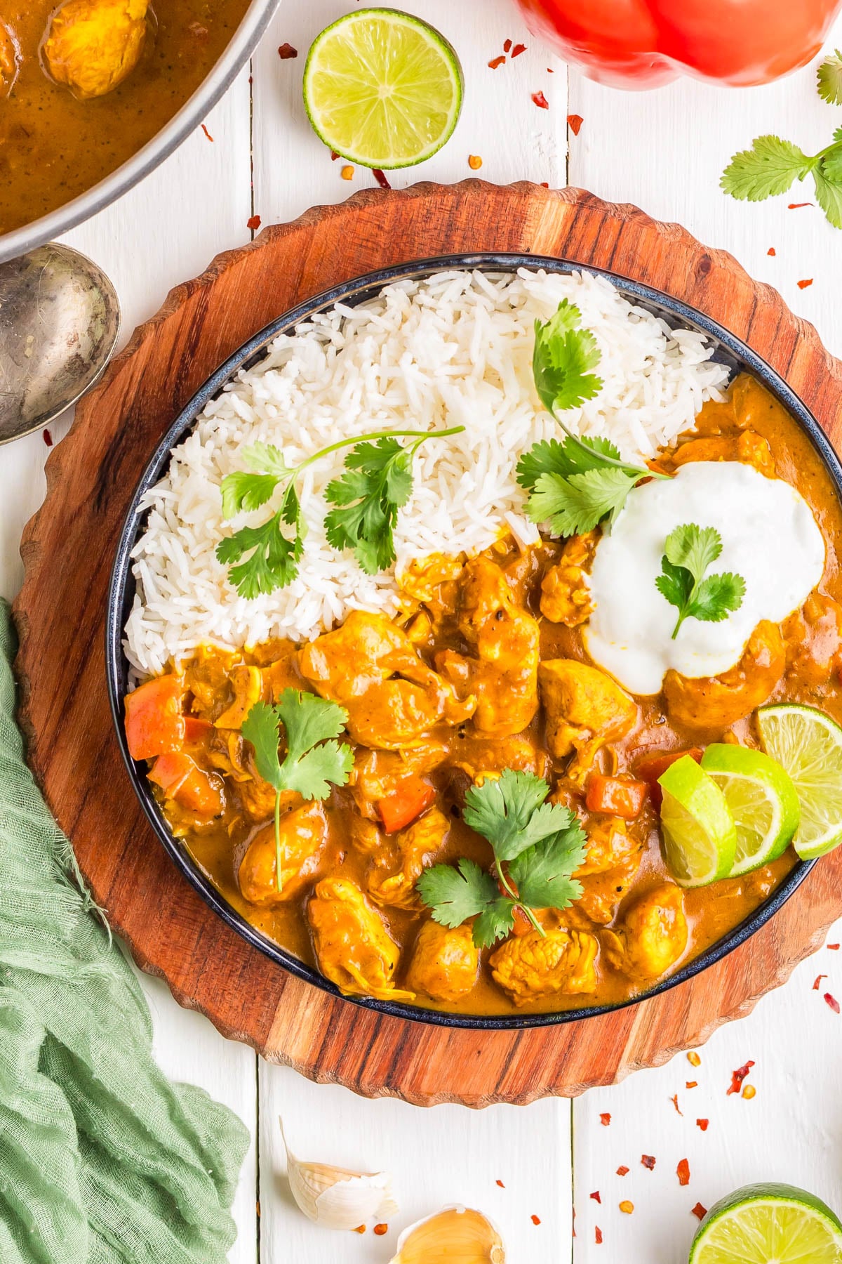 A bowl with white rice and chicken curry topped with cilantro and a dollop of yogurt, garnished with lime slices, on a wooden board.