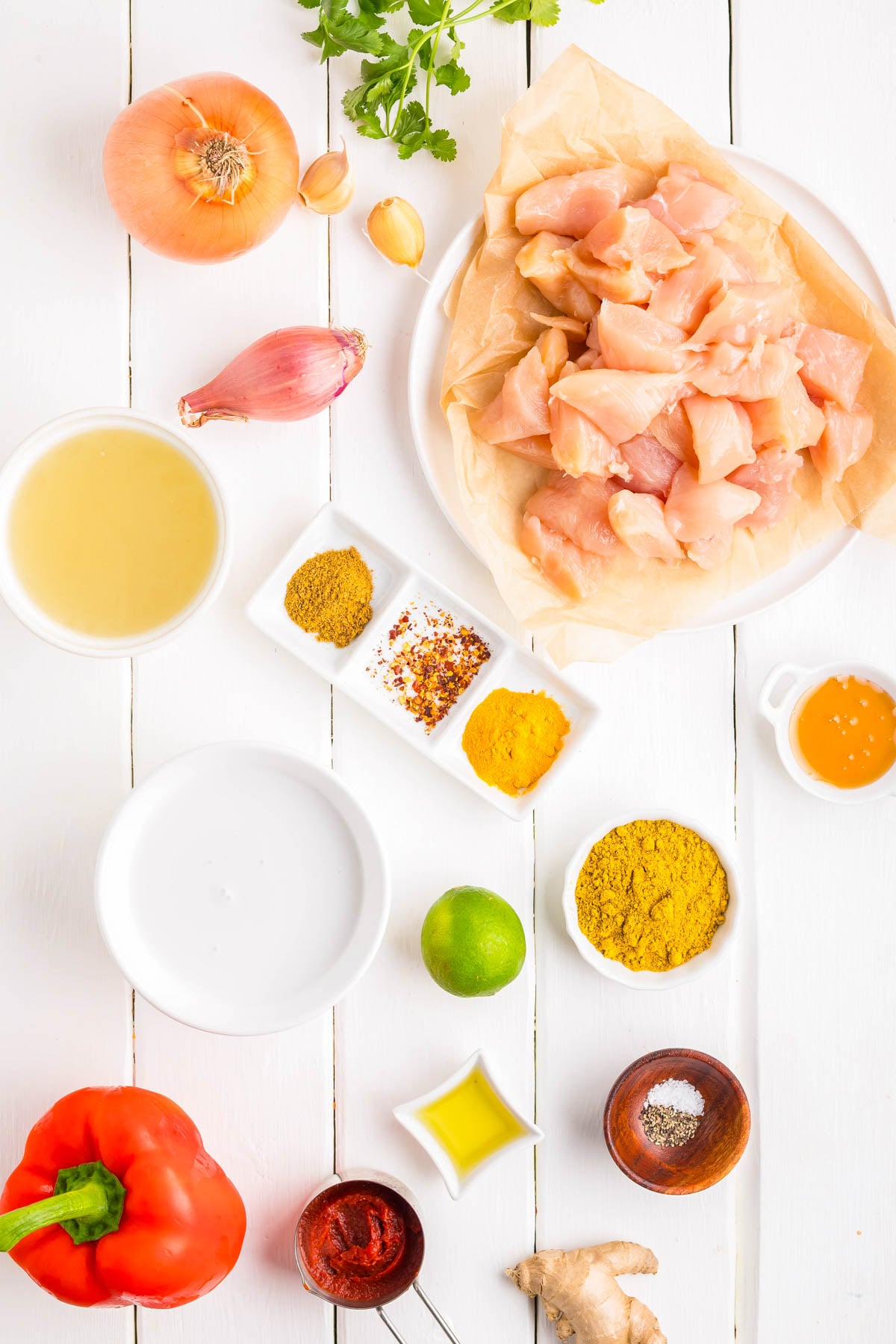 Overhead view of raw diced chicken, spices, red pepper, onion, garlic, shallot, lime, olive oil, ginger, broth, and other seasoning ingredients on a white table.