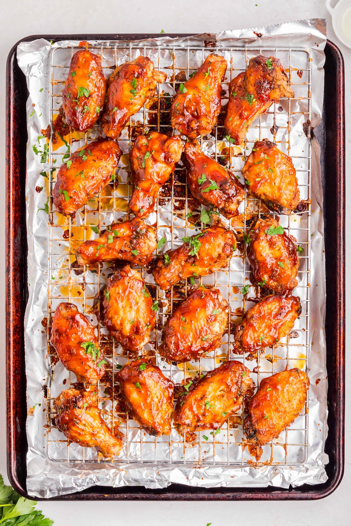 A tray of baked chicken wings coated in sauce, arranged on a metal rack over foil, garnished with chopped herbs.