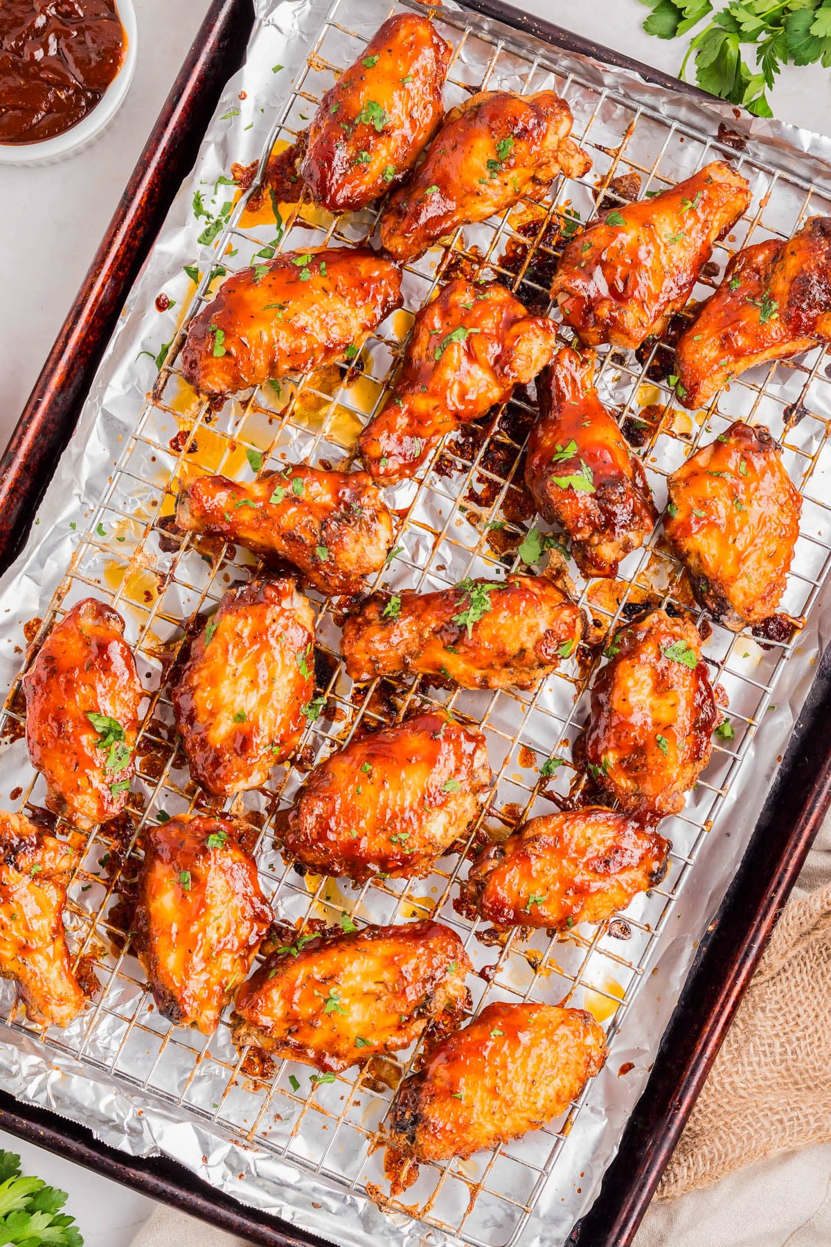Baked chicken wings coated in sauce are arranged on a wire rack over a foil-lined baking sheet, with a bowl of sauce and some herbs in the background.