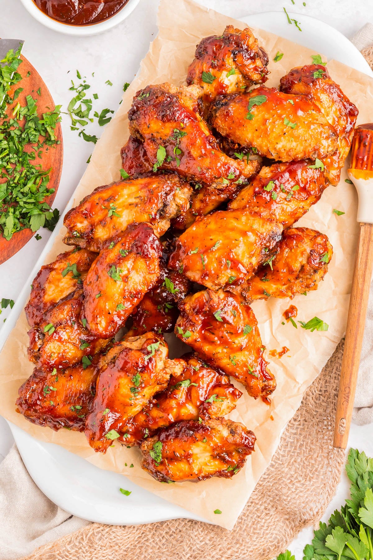 A platter of glazed chicken wings garnished with chopped herbs sits on parchment paper, with a bowl of sauce and chopped herbs nearby.