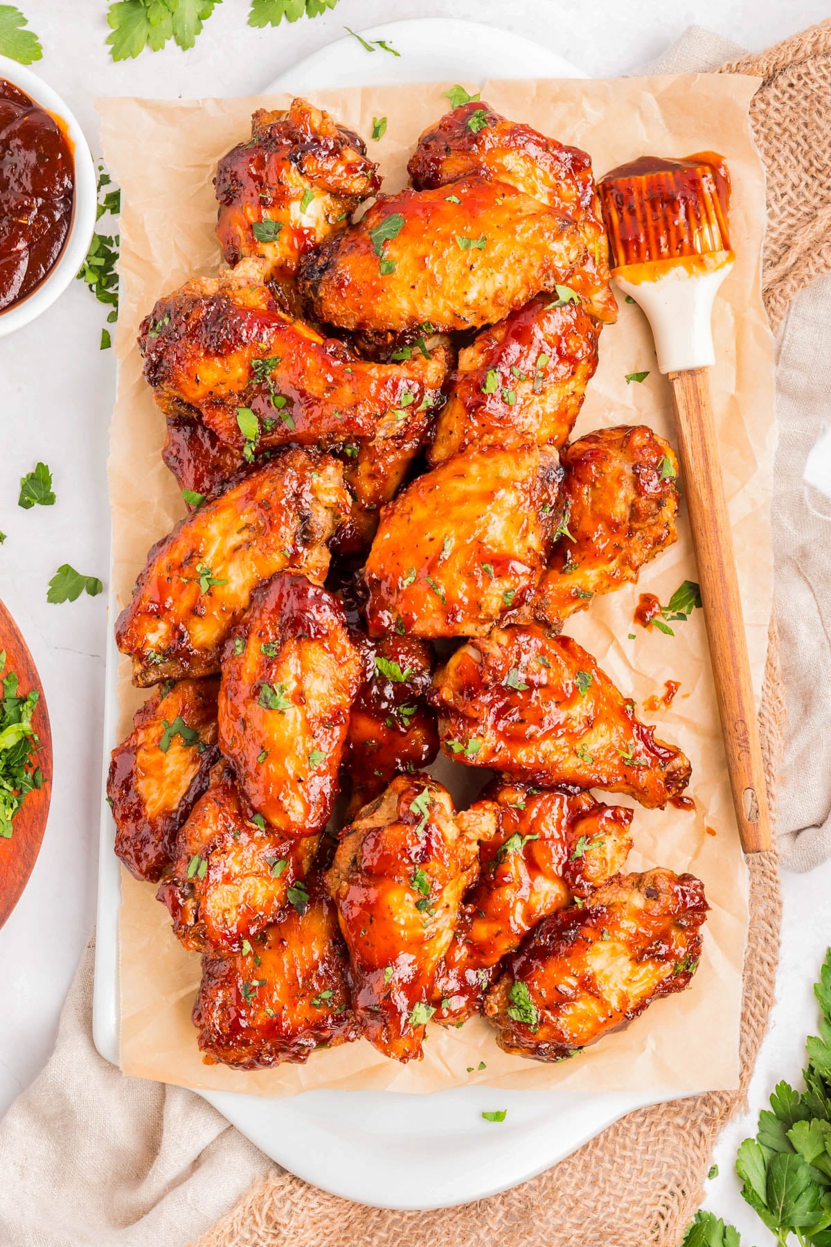 A pile of glazed chicken wings garnished with chopped herbs on parchment paper, with a basting brush and a bowl of sauce nearby.