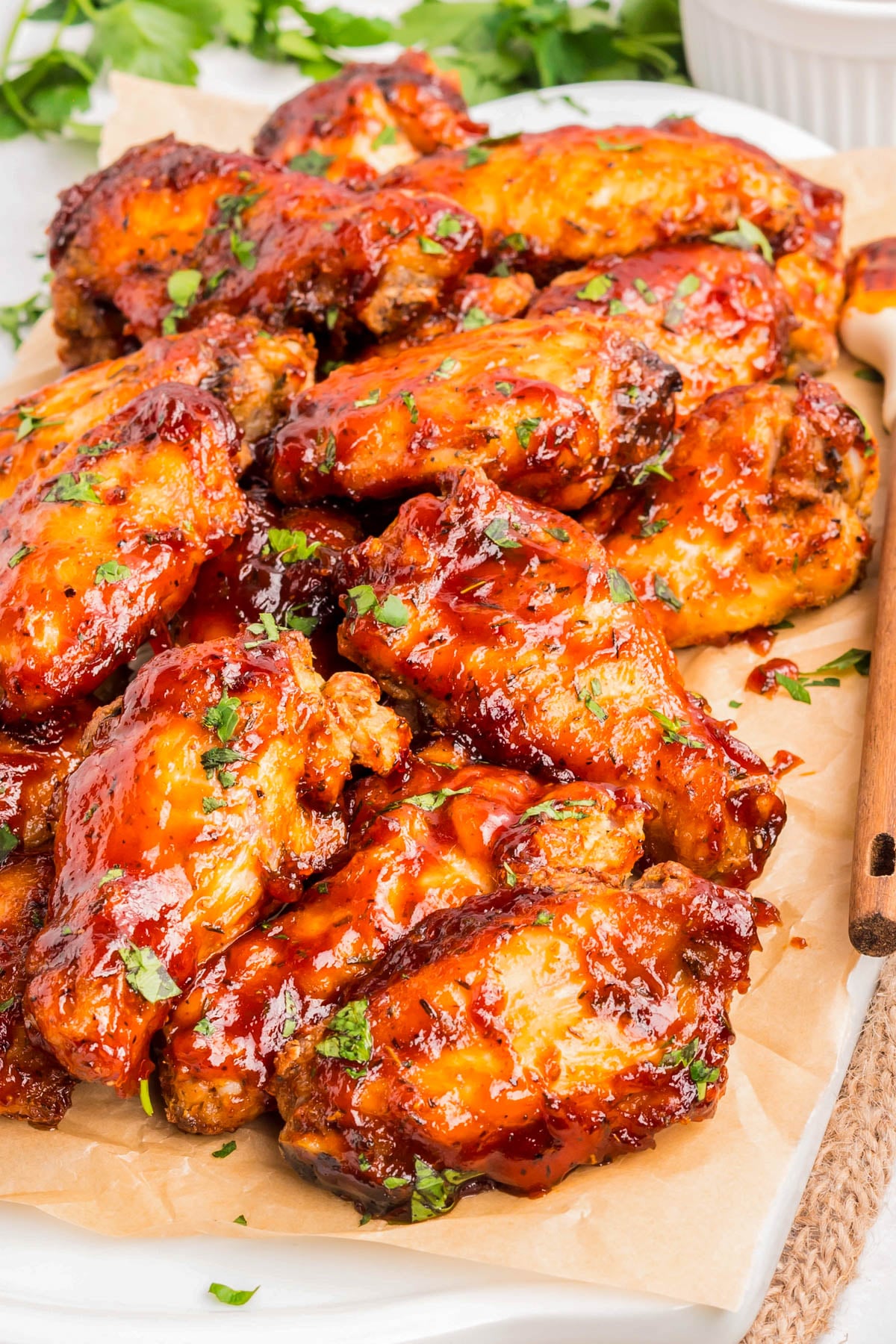 A platter of glazed chicken wings garnished with chopped herbs, arranged on parchment paper with a wooden serving utensil nearby.