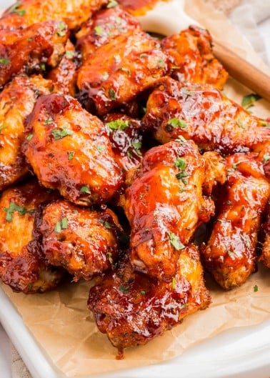 A tray of glazed chicken wings garnished with chopped herbs, placed on parchment paper, with a basting brush in the background.