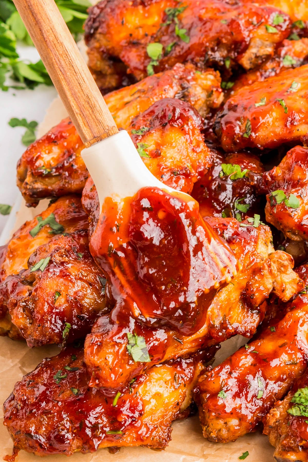 A brush spreads barbecue sauce over a pile of cooked chicken wings, garnished with chopped parsley.