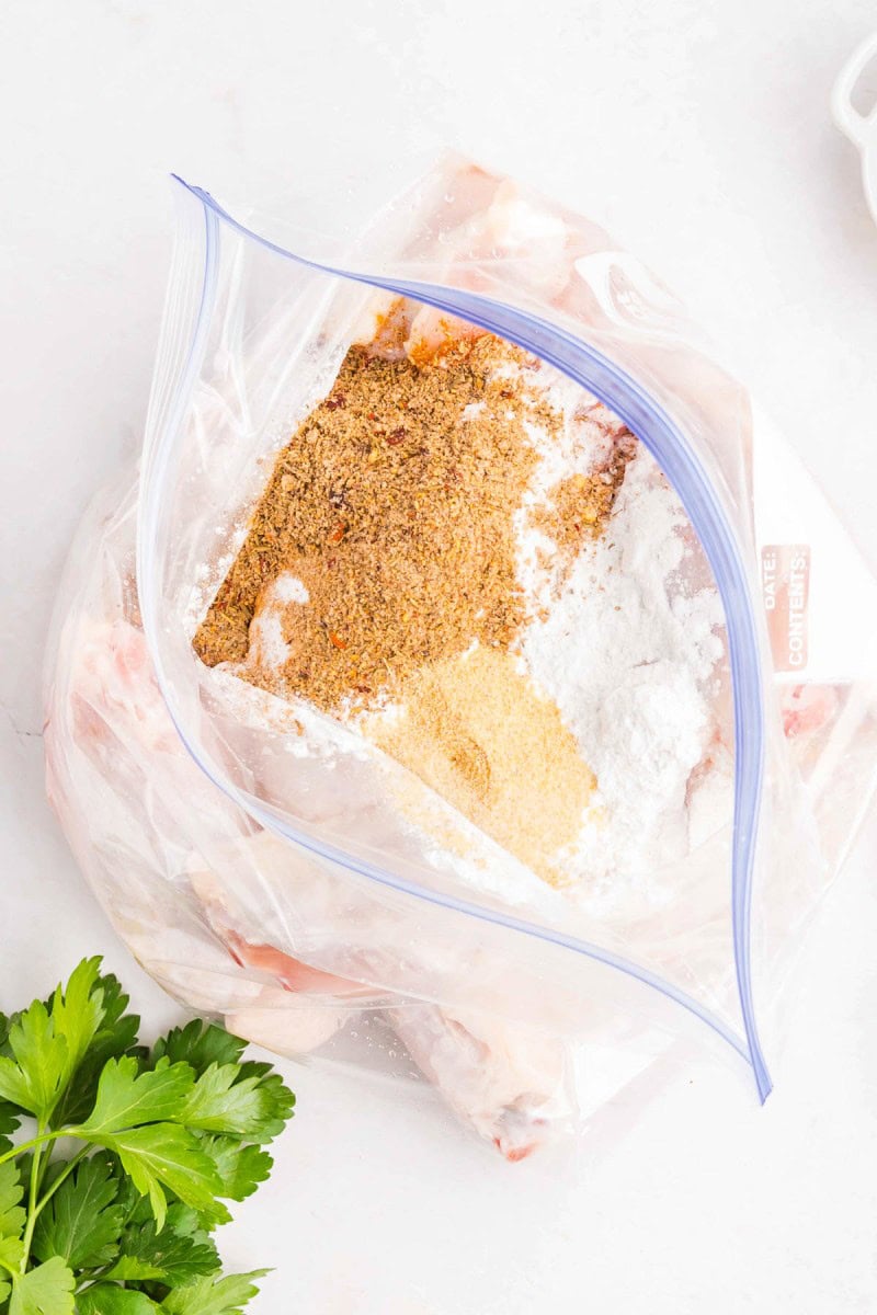 A plastic zip-top bag with raw chicken wings, flour, and various dry seasonings inside, on a white surface next to fresh parsley.