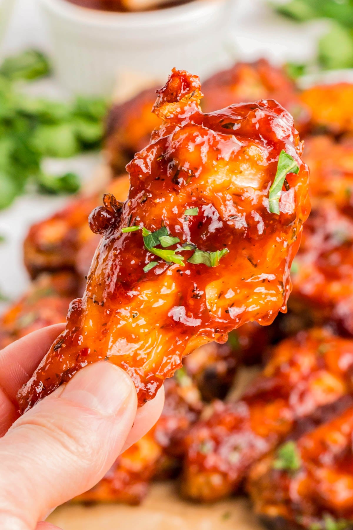 A hand holds a glazed chicken wing garnished with chopped herbs, with more sauced wings blurred in the background.