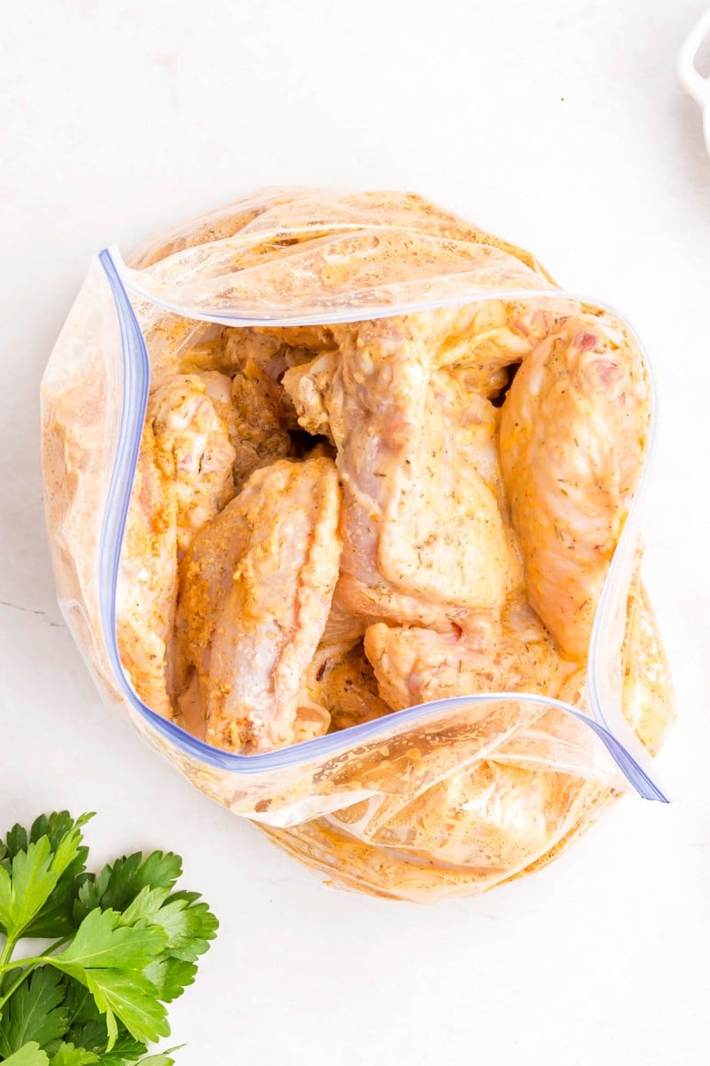 Raw chicken wings coated in marinade inside a partially open zip-top plastic bag on a white surface, with parsley leaves nearby.