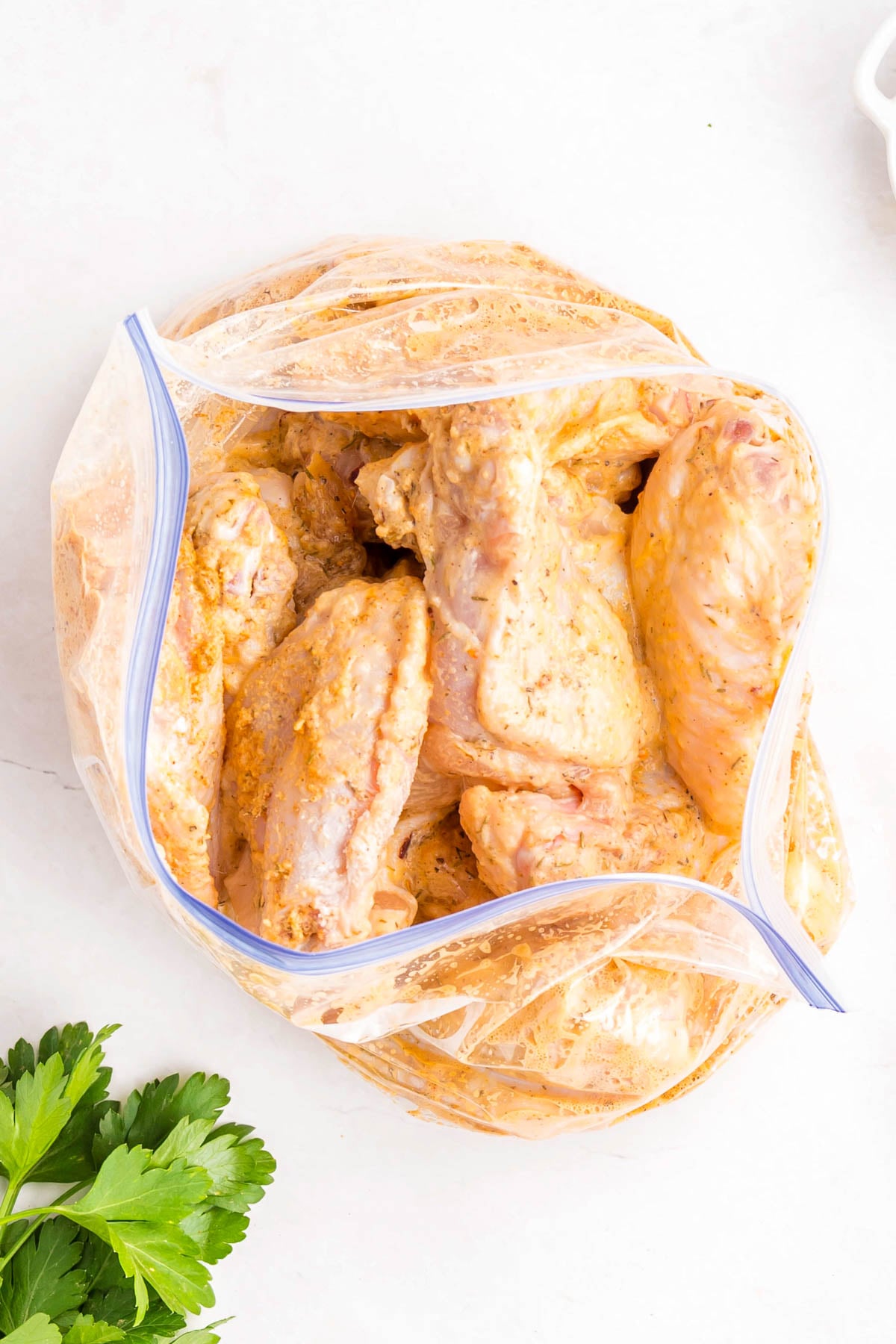 Raw chicken wings coated in marinade inside a partially open zip-top plastic bag on a white surface, with parsley leaves nearby.