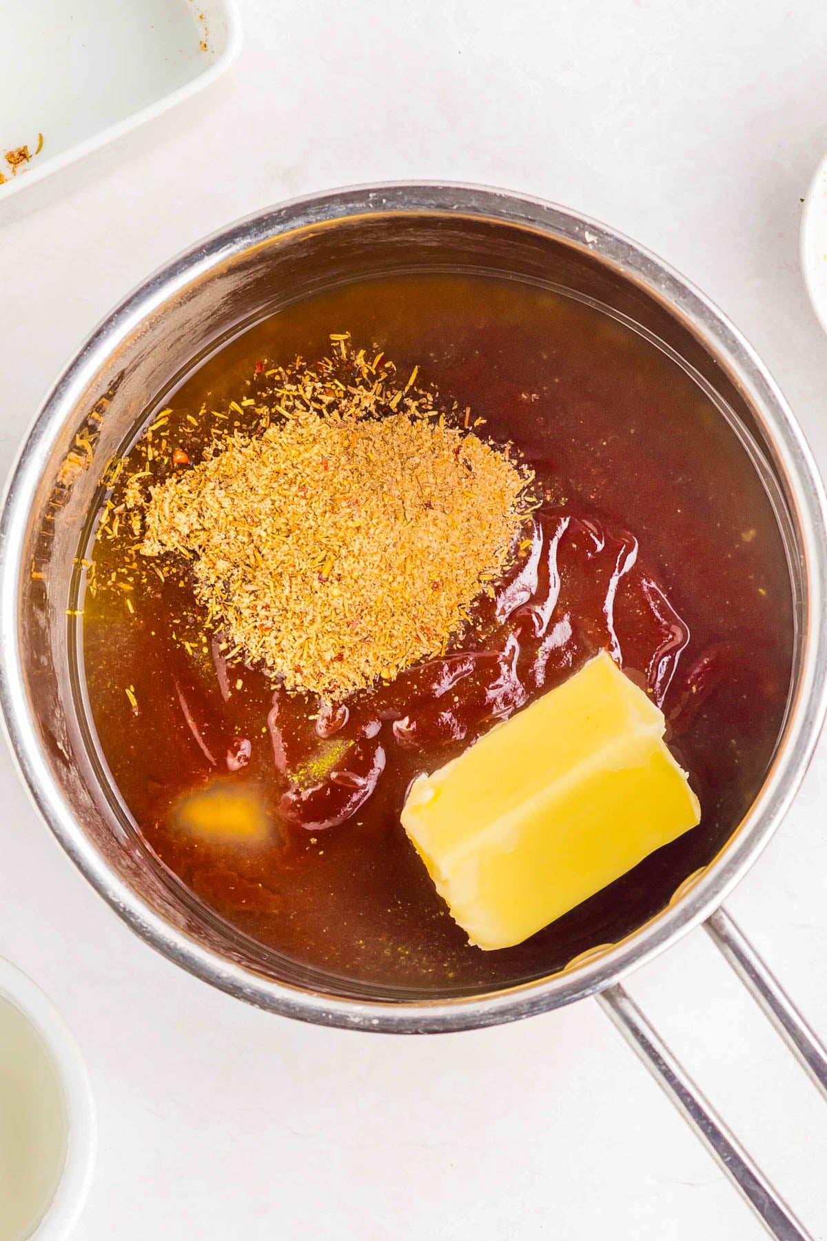 A saucepan contains a block of butter, seasoning, and a thick reddish sauce, possibly for mixing or cooking.