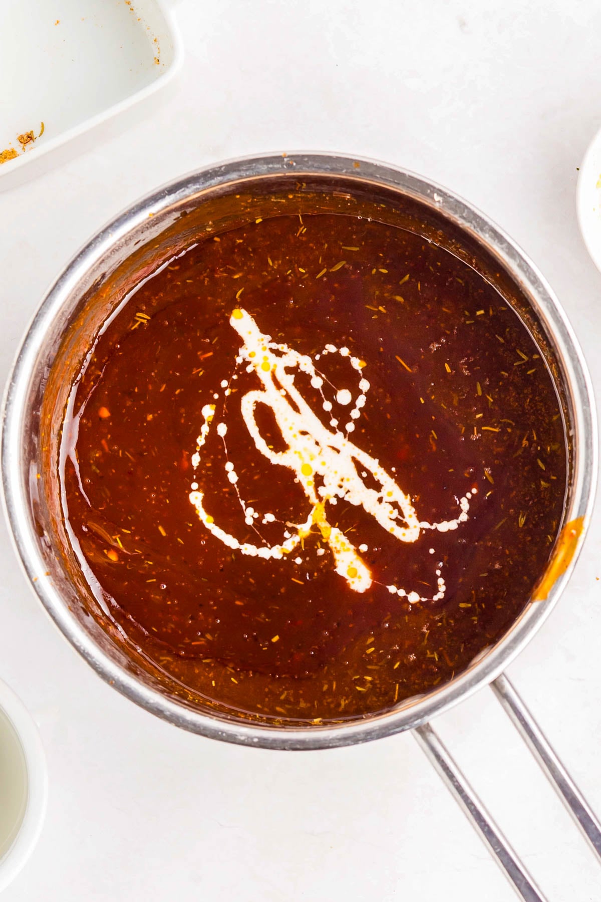 A metal saucepan filled with reddish-brown sauce, topped with a swirl of cream, sits on a white surface.