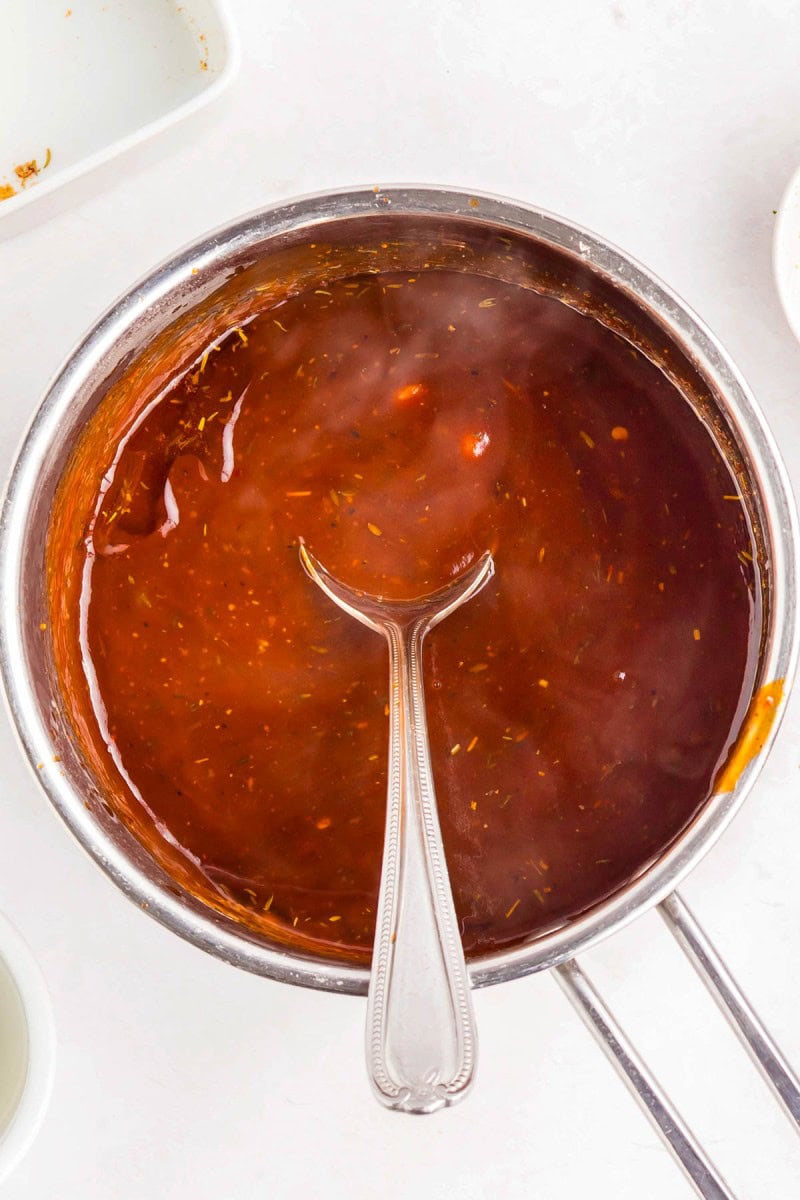 A stainless steel saucepan filled with reddish-brown barbecue sauce, with a metal spoon resting inside.