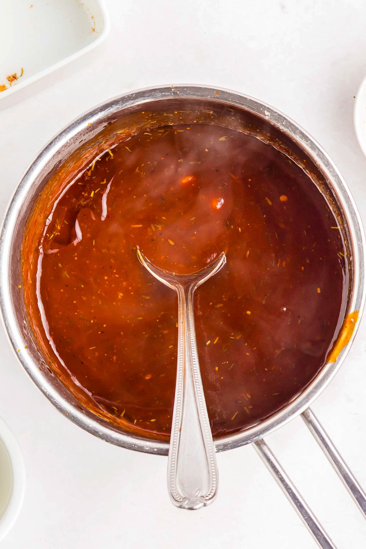 A stainless steel saucepan filled with reddish-brown barbecue sauce, with a metal spoon resting inside.