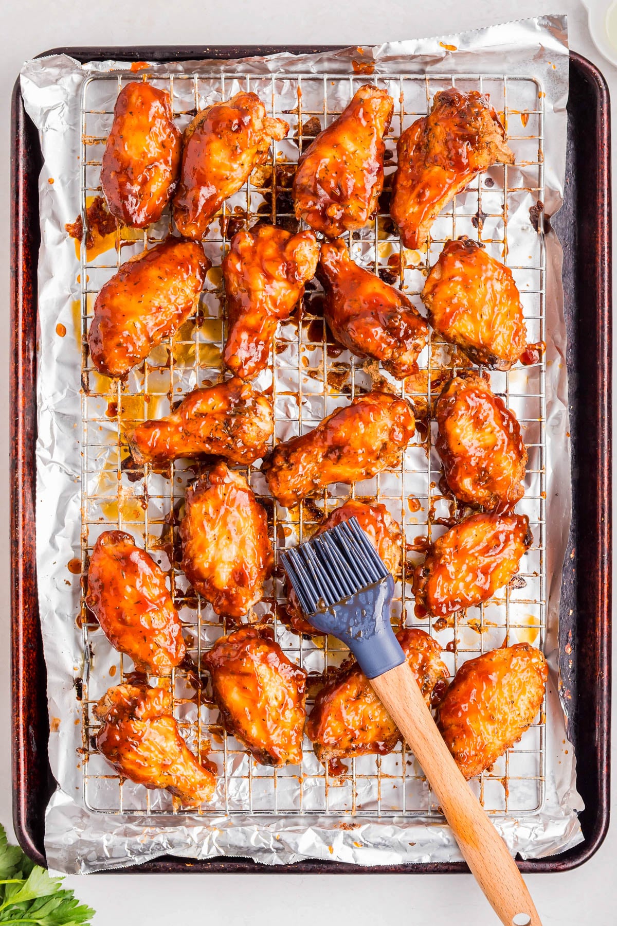 Baked chicken wings coated in sauce are arranged on a wire rack over a foil-lined baking sheet, with a basting brush resting on top.