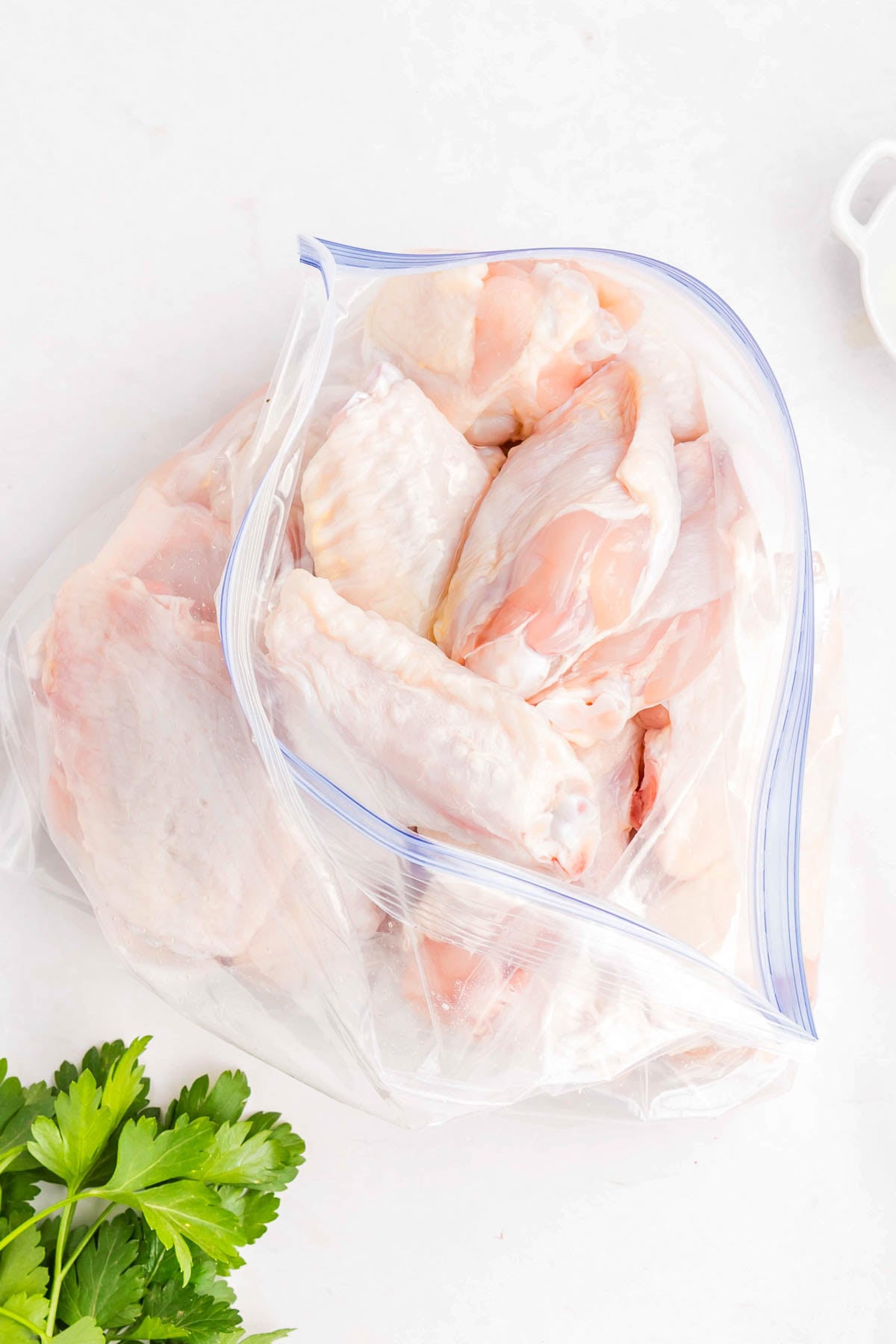 Raw chicken wings in a clear resealable plastic bag on a white surface, with fresh parsley nearby.