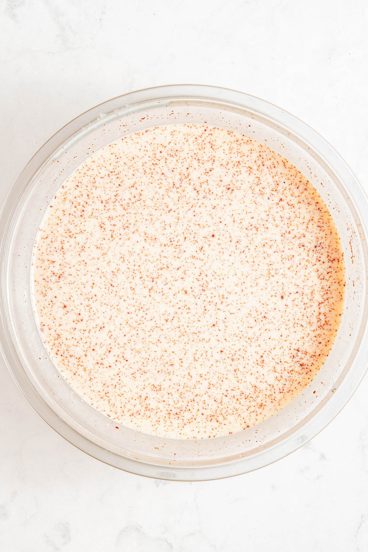 A glass bowl filled with a creamy, speckled liquid mixture sits on a white marble surface.